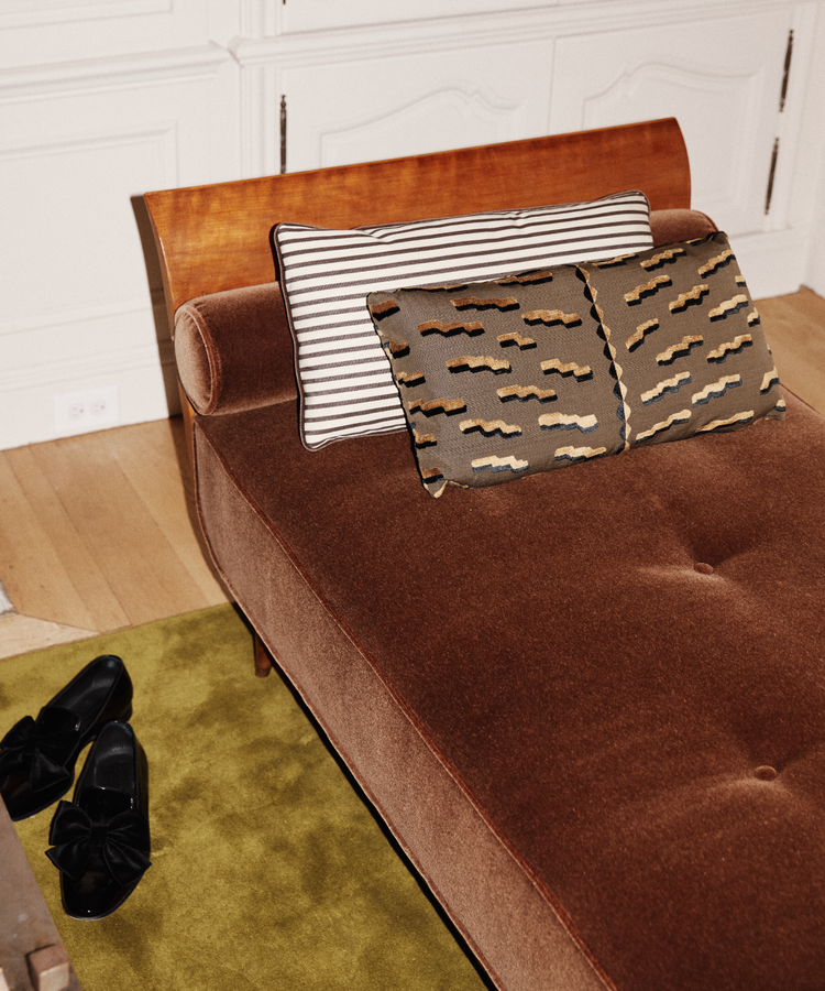 A brown upholstered chaise lounge with Polo Lumbar Cushion, black shoes on an olive green rug, and white cabinetry in the background.