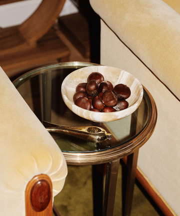 A Nutcracker sits beside a bowl of whole chestnuts on a round glass side table between two beige upholstered chairs.