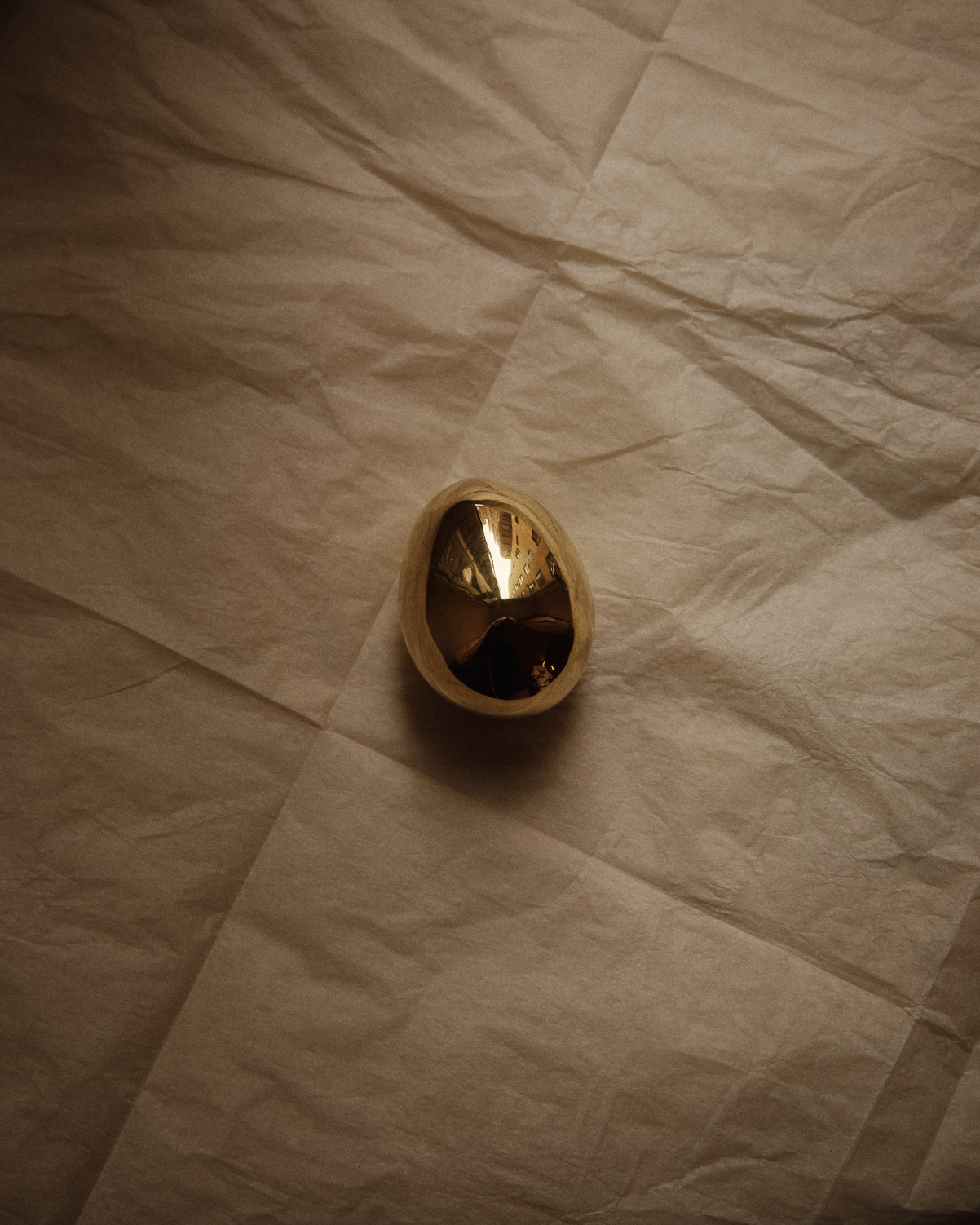 The Egg Paperweight, a reflective polished brass desk accessory partially encased in a light shell, sits on crumpled beige paper—evoking the iconic style of the Auböck workshop.