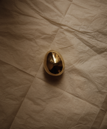The Egg Paperweight, a reflective polished brass desk accessory partially encased in a light shell, sits on crumpled beige paper—evoking the iconic style of the Auböck workshop.