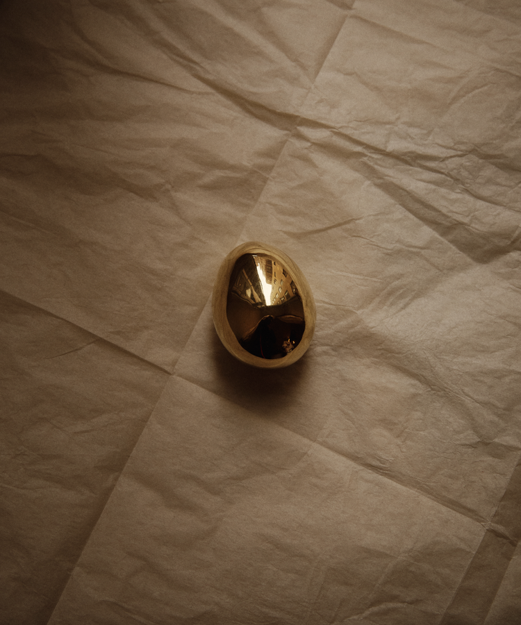 The Egg Paperweight, a reflective polished brass desk accessory partially encased in a light shell, sits on crumpled beige paper—evoking the iconic style of the Auböck workshop.