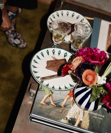 A Vito Small Plate with a slice of chocolate cake, two clear drinks in Vito glasses, a handmade flower vase, and a photo book rest on a table; part of a person's legs can be seen in the background.