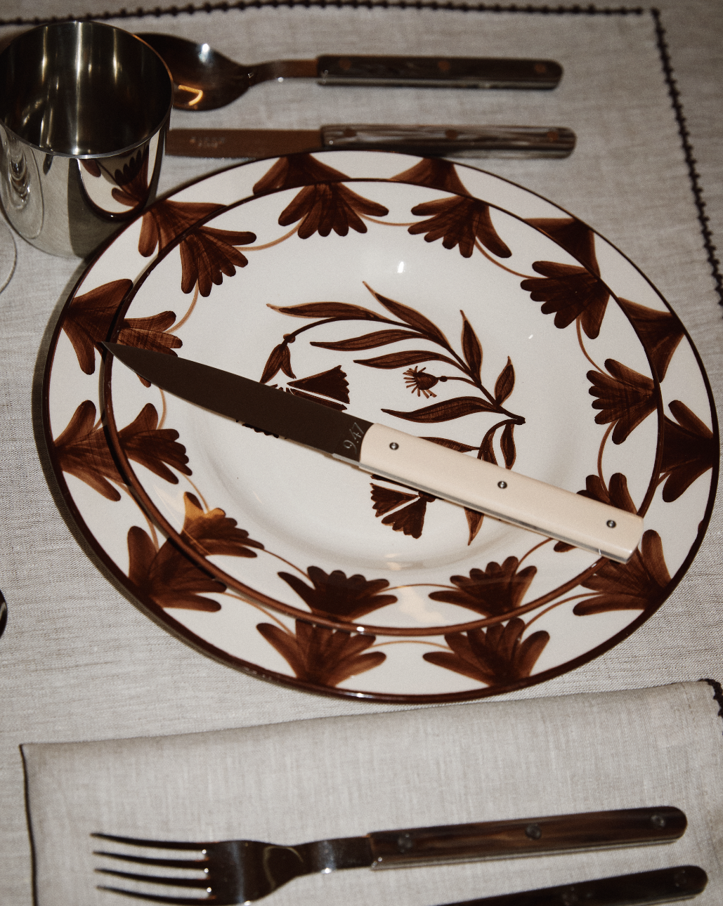 A ceramic plate with brown floral patterns is set with a 9.47 Table Knife across it, alongside a fork, spoon, metal cup, and folded napkin on a linen placemat—a true showcase of French craftsmanship.