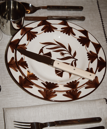A ceramic plate with brown floral patterns is set with a 9.47 Table Knife across it, alongside a fork, spoon, metal cup, and folded napkin on a linen placemat—a true showcase of French craftsmanship.