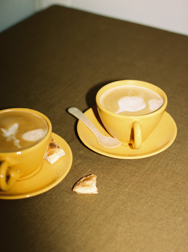 Two yellow Guido Coffee Set cups and saucers filled with coffee and foam art sit on a brown table beside a small spoon and pastry pieces.