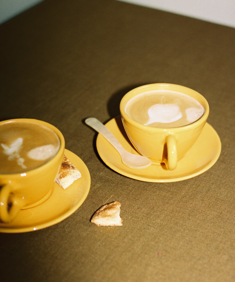 Two yellow Guido Coffee Set cups and saucers filled with coffee and foam art sit on a brown table beside a small spoon and pastry pieces.