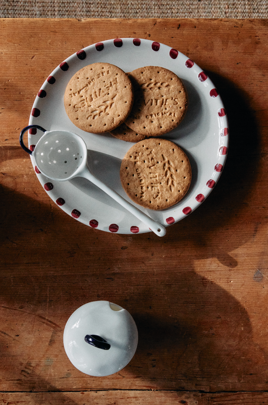 Three round biscuits rest on the Nina Small Plate, Set of 4, featuring hand-glazed ceramic with artisanal detail. The plate sits on a wooden table beside a white strainer and a small white container with a dark handle.