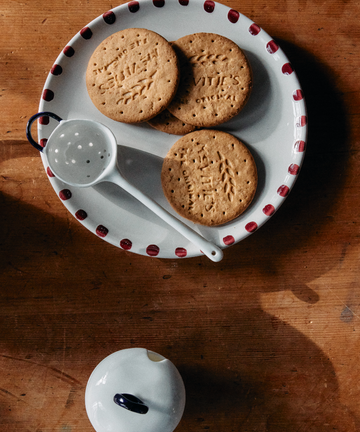 Three round biscuits rest on the Nina Small Plate, Set of 4, featuring hand-glazed ceramic with artisanal detail. The plate sits on a wooden table beside a white strainer and a small white container with a dark handle.
