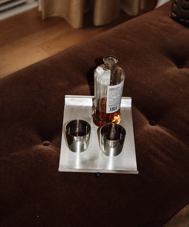 The Alexander Stemless Tumbler Set of 2 sits on a rectangular tray with a bottle of liquor atop a brown cushioned surface, lending a modern touch to the tableware arrangement.
