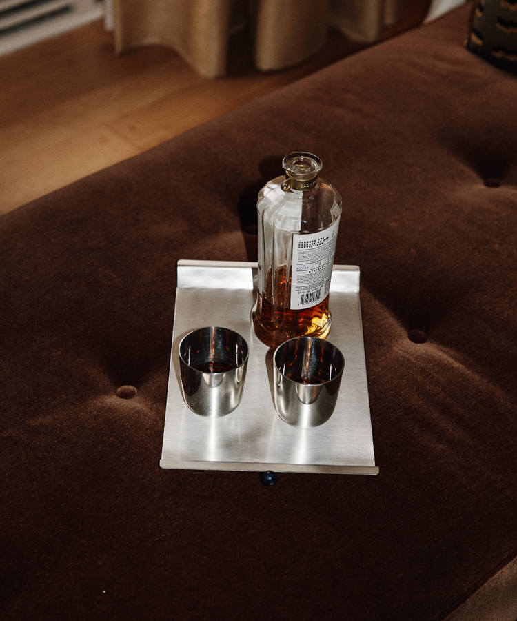 The Alexander Stemless Tumbler Set of 2 sits on a rectangular tray with a bottle of liquor atop a brown cushioned surface, lending a modern touch to the tableware arrangement.