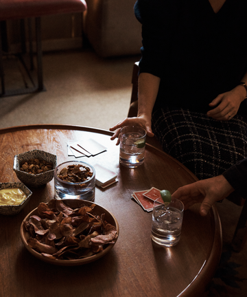 Two people sit at a round wooden table with snacks, a deck of cards, and two Boris Tumblers filled with water and lime. One person reaches for their drink.