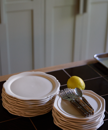 Two stacks of white creamware plates, featuring the Cirrus Octagonal Plate, sit on a kitchen counter with forks and spoons placed on one stack and a lemon nearby.