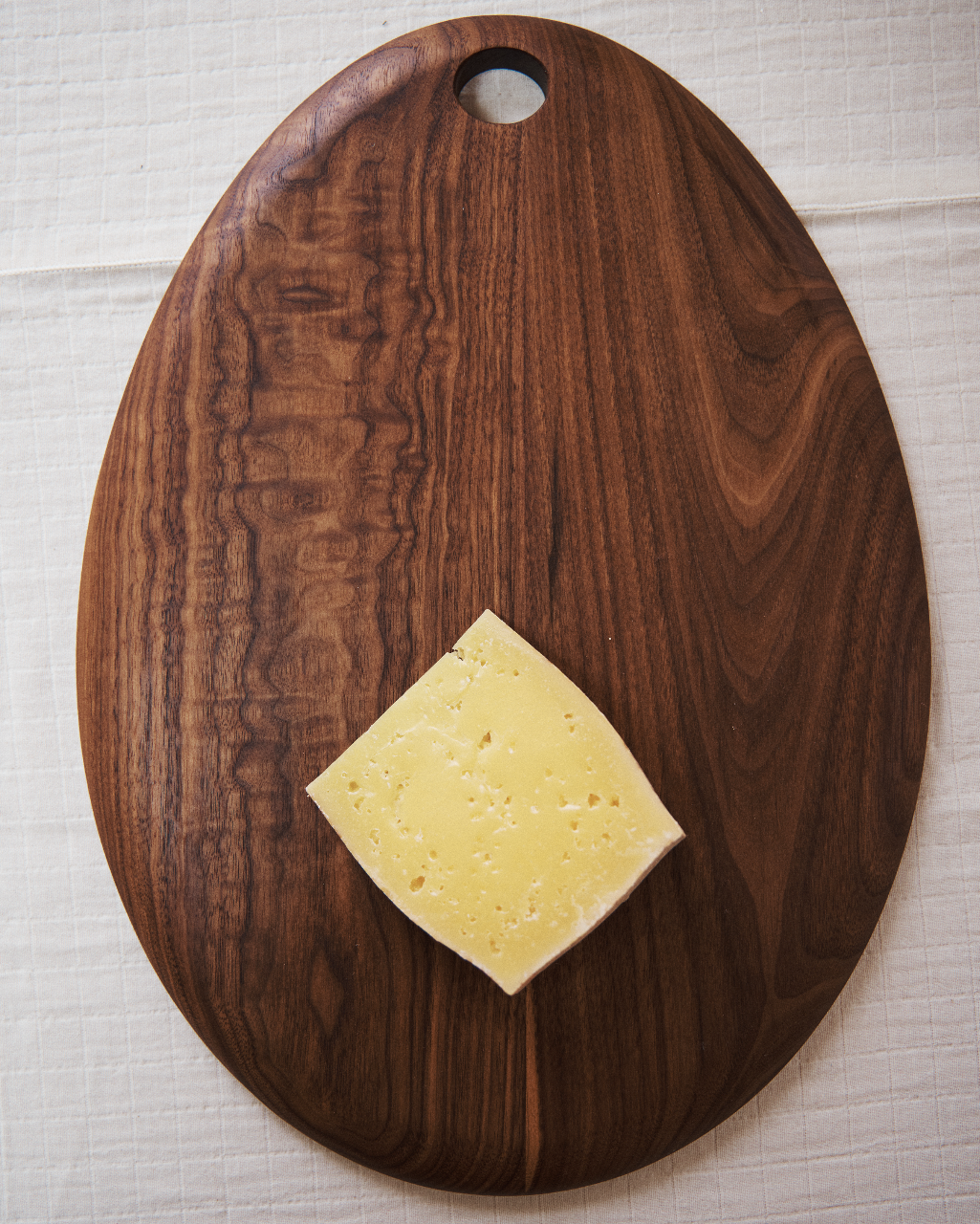 A single slice of yellow cheese with holes is placed on an Alden Egg Board, a dark walnut board featuring a rounded shape and a hole at the top.