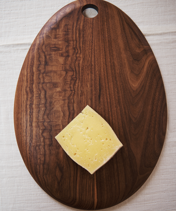 A single slice of yellow cheese with holes is placed on an Alden Egg Board, a dark walnut board featuring a rounded shape and a hole at the top.