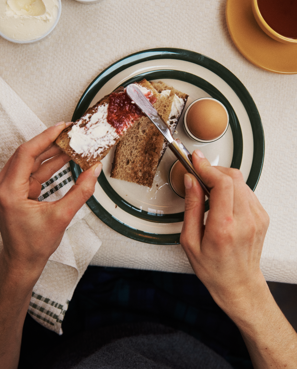 A person spreads jam on bread with cream cheese beside two soft-boiled eggs, using a Butter Knife in the kitchen, with a cup and a bowl visible in the background.