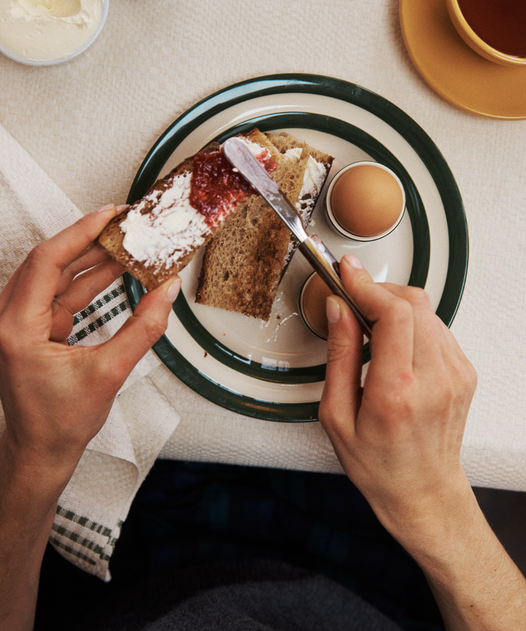 A person spreads jam on bread with cream cheese beside two soft-boiled eggs, using a Butter Knife in the kitchen, with a cup and a bowl visible in the background.