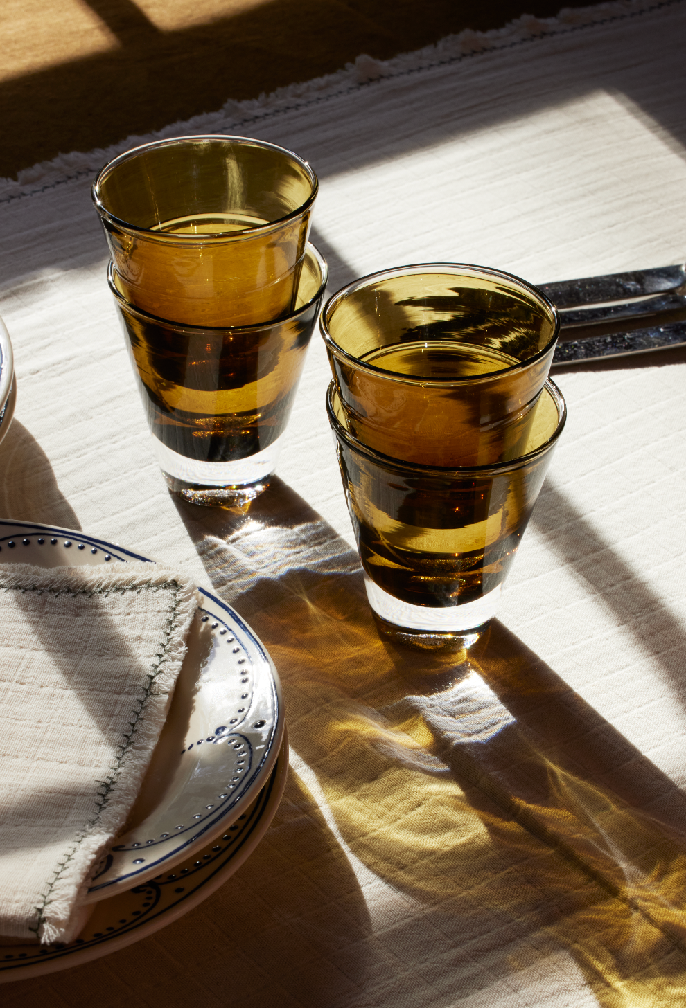 Two Gem Glasses, set of 2, in amber are stacked together on a white tablecloth beside plates, a folded napkin, and cutlery in sunlight—capturing the relaxed elegance of the South of France.