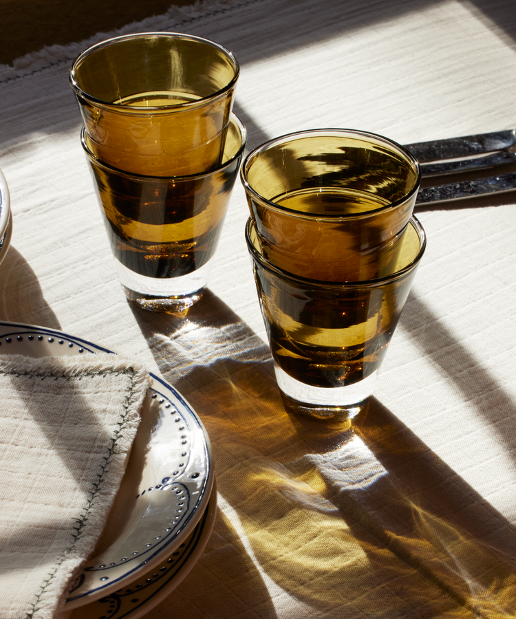Two Gem Glasses, set of 2, in amber are stacked together on a white tablecloth beside plates, a folded napkin, and cutlery in sunlight—capturing the relaxed elegance of the South of France.