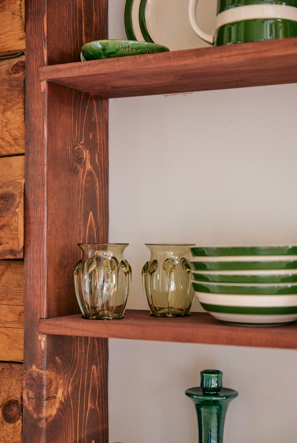 A wooden shelf displays green and white striped ceramic bowls, clear glass tumblers, and the Carlotta Vase in mouth-blown green glass, showcasing Bohemian artistry against a crisp white wall.