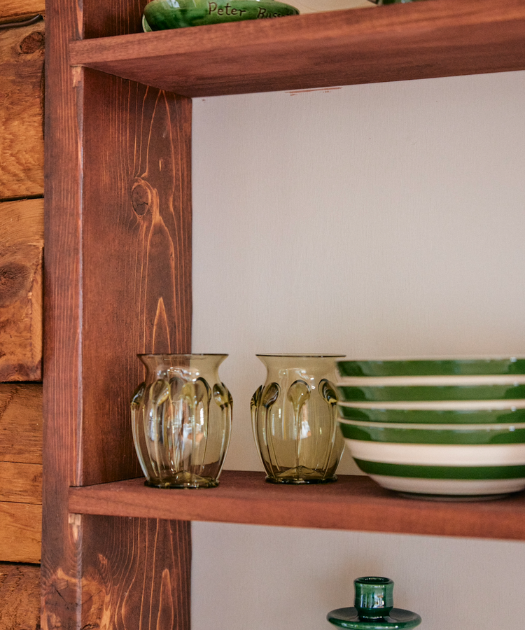 A wooden shelf displays green and white striped ceramic bowls, clear glass tumblers, and the Carlotta Vase in mouth-blown green glass, showcasing Bohemian artistry against a crisp white wall.