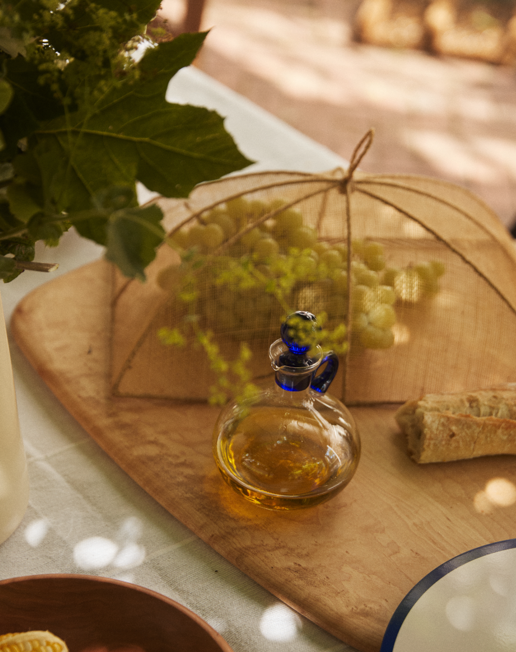 A glass bottle of olive oil, a piece of baguette, and grapes are placed under the Jacqueline Foodcover on a wooden board resting on a table.