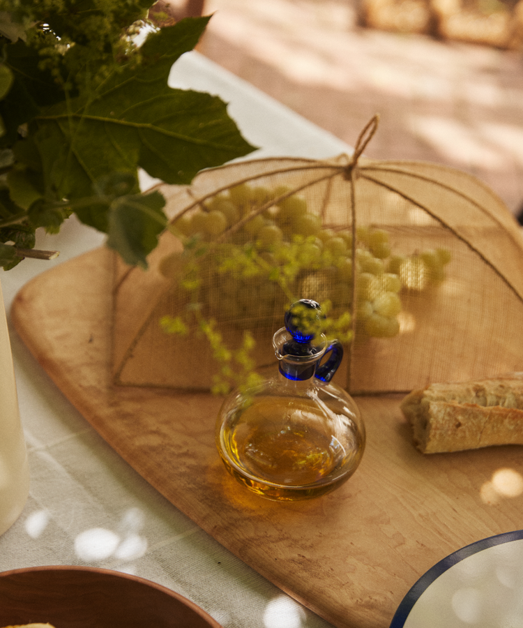 A glass bottle of olive oil, a piece of baguette, and grapes are placed under the Jacqueline Foodcover on a wooden board resting on a table.
