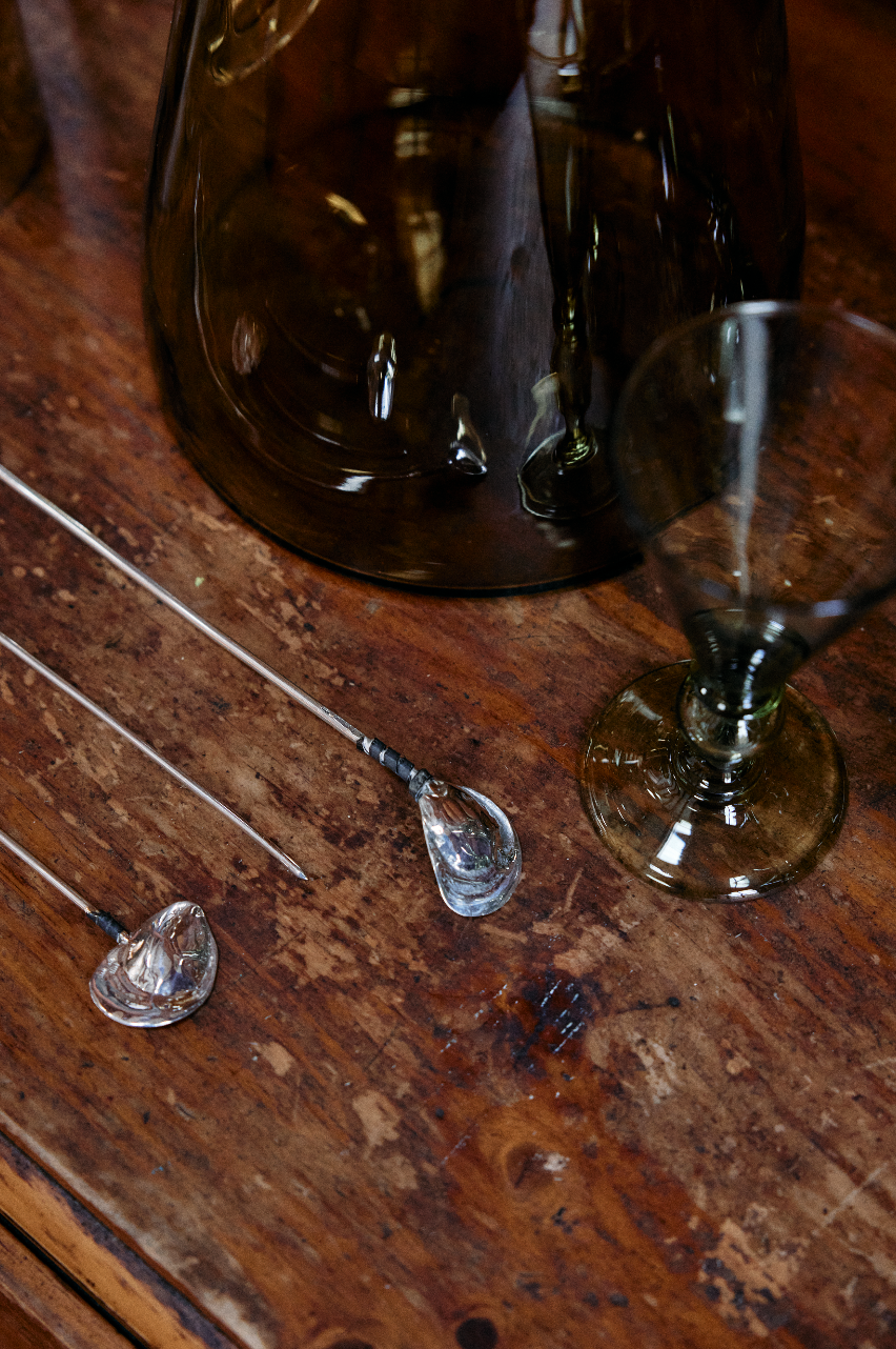 An Oyster Shell Cocktail Stirrer rests on a wooden surface alongside a tinted glass goblet and a large glass container.