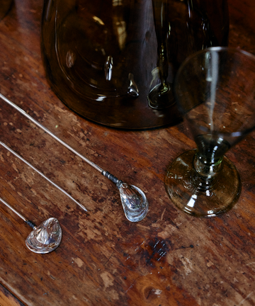 An Oyster Shell Cocktail Stirrer rests on a wooden surface alongside a tinted glass goblet and a large glass container.