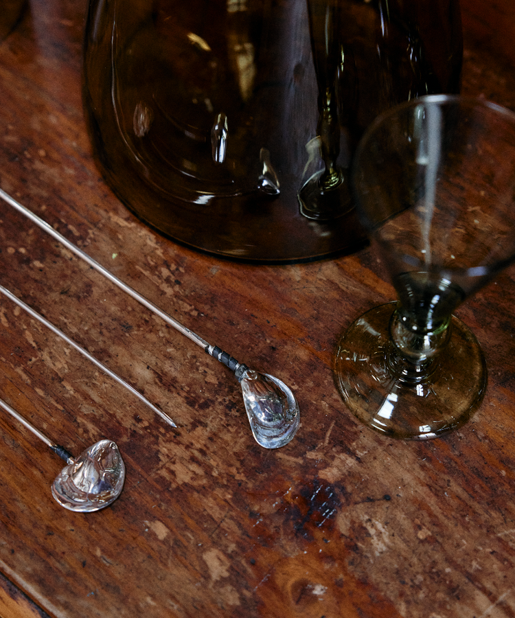 An Oyster Shell Cocktail Stirrer rests on a wooden surface alongside a tinted glass goblet and a large glass container.