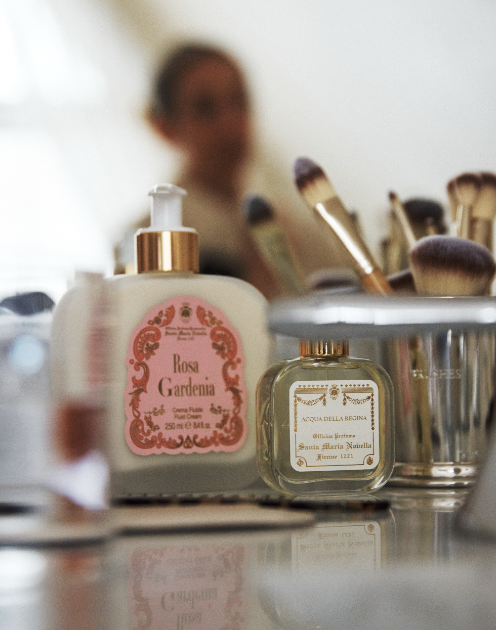 A bottle of Rosa Gardenia Fluid Body Cream and a glass perfume bottle rest on a vanity with makeup brushes, while a blurred figure in the background creates an elegant scene inspired by Florentine craftsmanship.