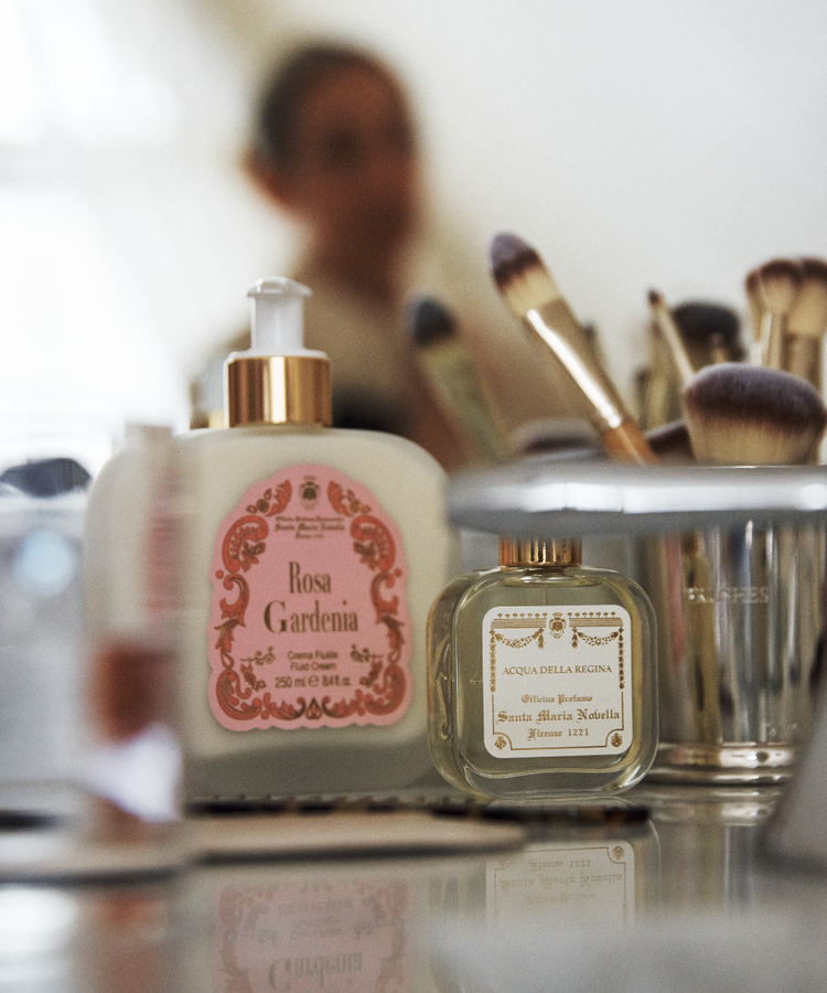 A bottle of Rosa Gardenia Fluid Body Cream and a glass perfume bottle rest on a vanity with makeup brushes, while a blurred figure in the background creates an elegant scene inspired by Florentine craftsmanship.