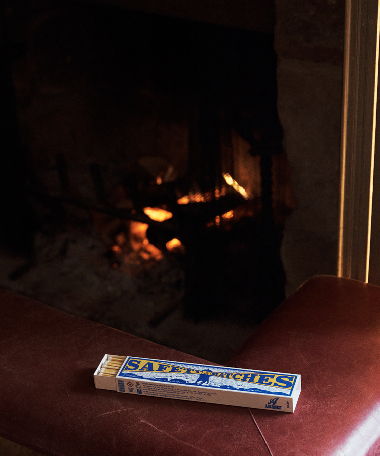 A box of Luxury Matches, filled with extra-long matches, rests on a brown leather surface in front of a lit fireplace with a warm, glowing fire.