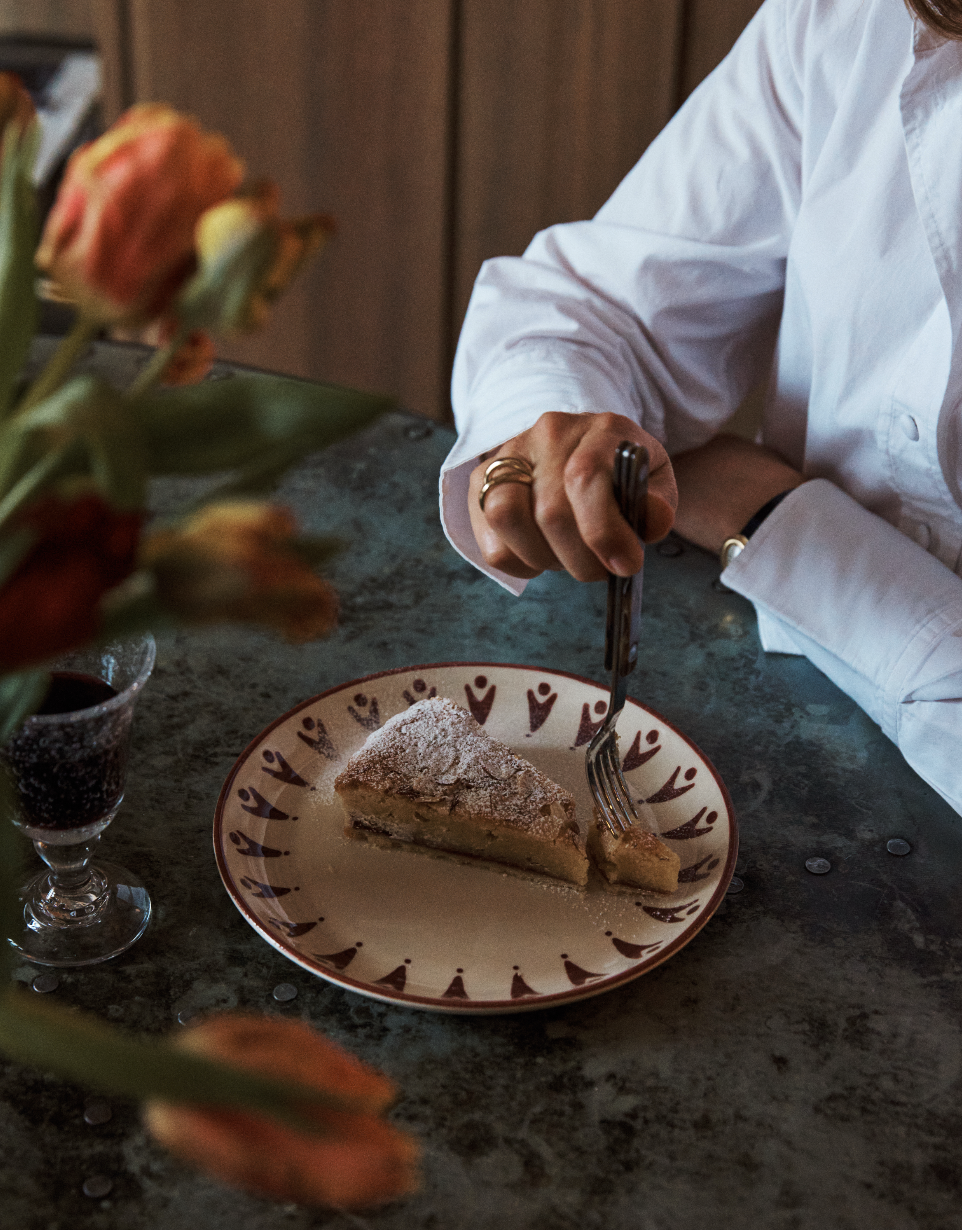A person in a white shirt holds a fork poised over a slice of cake on a decorated handmade plate from the Vito Small Plate, Set of 4, with a glass and some flowers nearby on the table—perfect for everyday use.