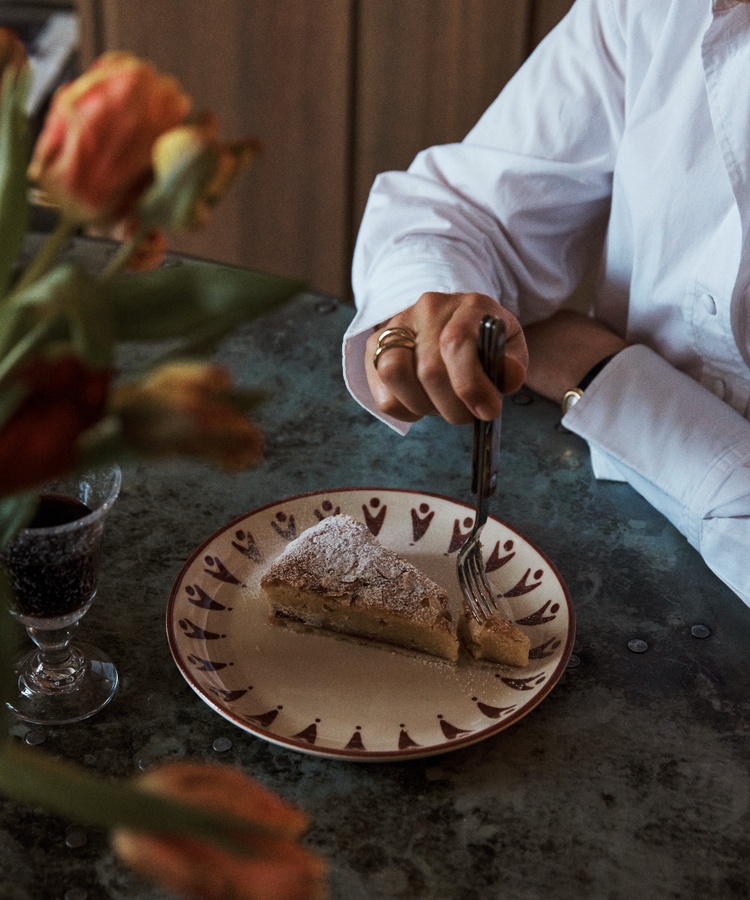 A person in a white shirt holds a fork poised over a slice of cake on a decorated handmade plate from the Vito Small Plate, Set of 4, with a glass and some flowers nearby on the table—perfect for everyday use.