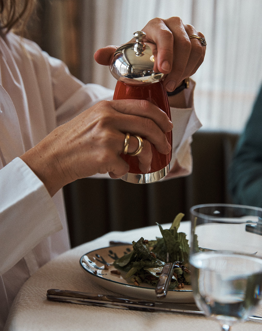A person in a white shirt uses the Giorgio Pepper Mill, known for its superior grinding mechanism, to season a salad on a plate at a set dining table with cutlery and a glass of water.