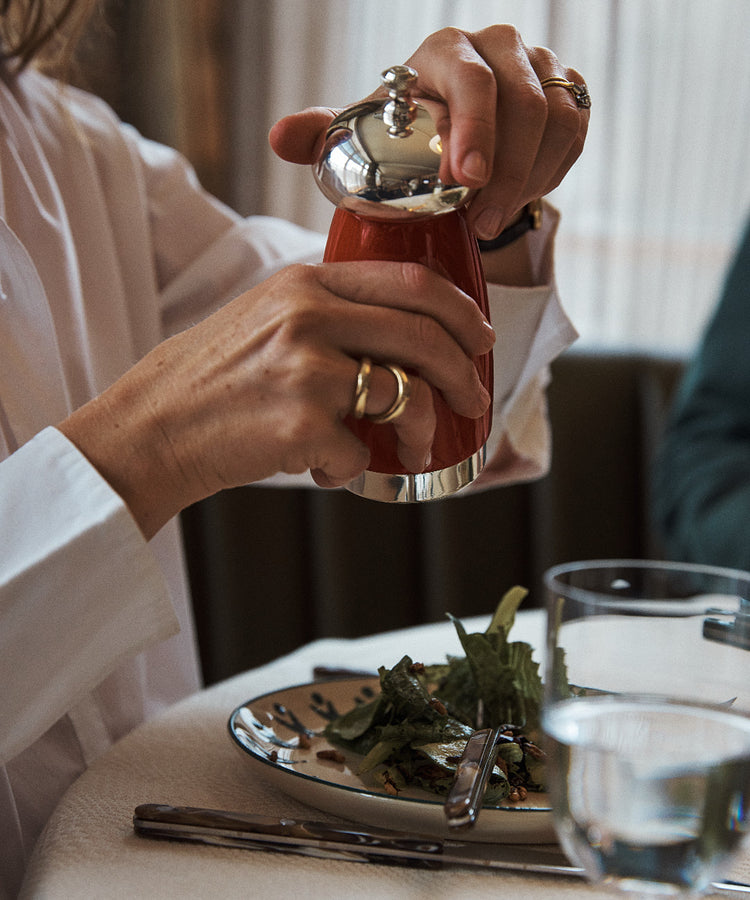 A person in a white shirt uses the Giorgio Pepper Mill, known for its superior grinding mechanism, to season a salad on a plate at a set dining table with cutlery and a glass of water.