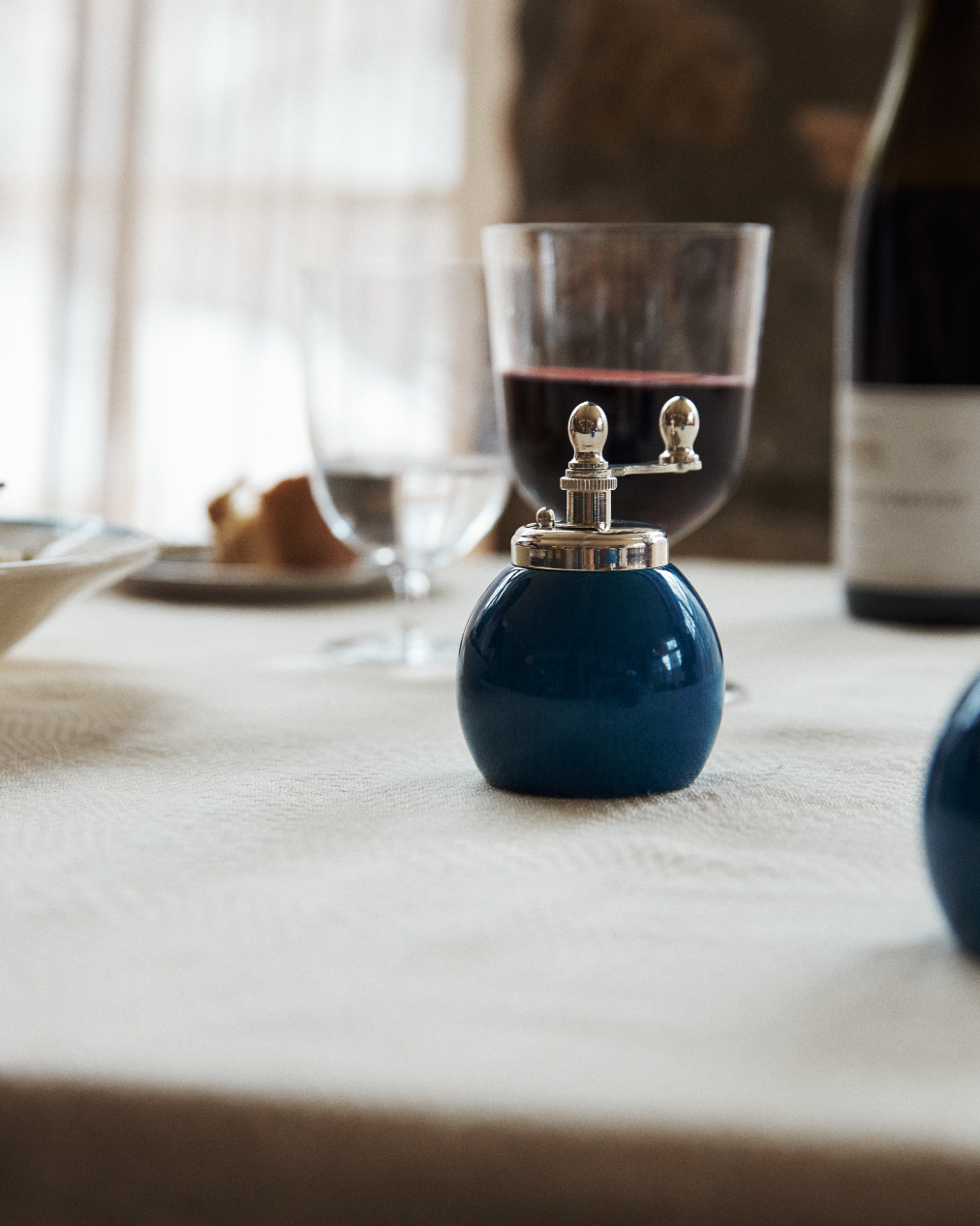 The Theo Pepper Mill and Salt Shaker, featuring a classic Italian design in blue, sits on a white tablecloth next to a wine glass, water glass, and a wine bottle.