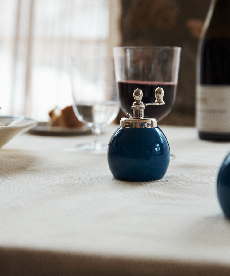The Theo Pepper Mill and Salt Shaker, featuring a classic Italian design in blue, sits on a white tablecloth next to a wine glass, water glass, and a wine bottle.