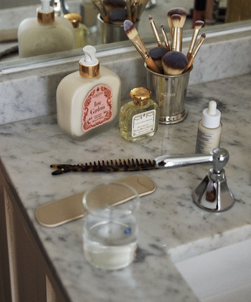A marble bathroom counter with a soap dispenser, perfume bottle, makeup brushes in a holder, serum dropper bottle, hair detangler, the Capri Sardine Comb, nail file, and a glass of water near a faucet.