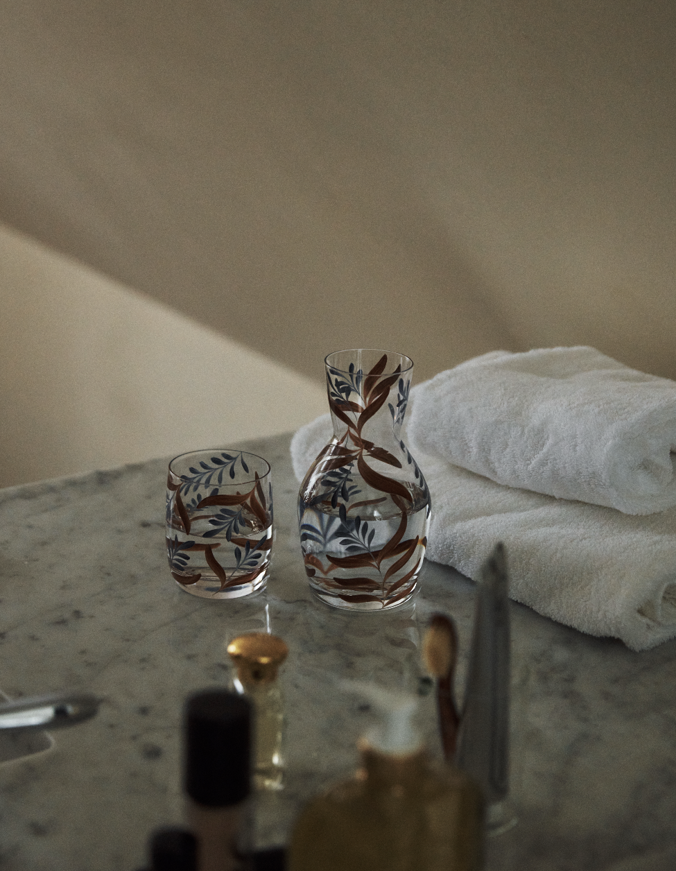 The Botanical Bedside Carafe and Glass Set, featuring a hand-painted botanical design, rests on a marble surface alongside two folded white towels and assorted toiletries.