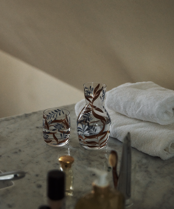 The Botanical Bedside Carafe and Glass Set, featuring a hand-painted botanical design, rests on a marble surface alongside two folded white towels and assorted toiletries.