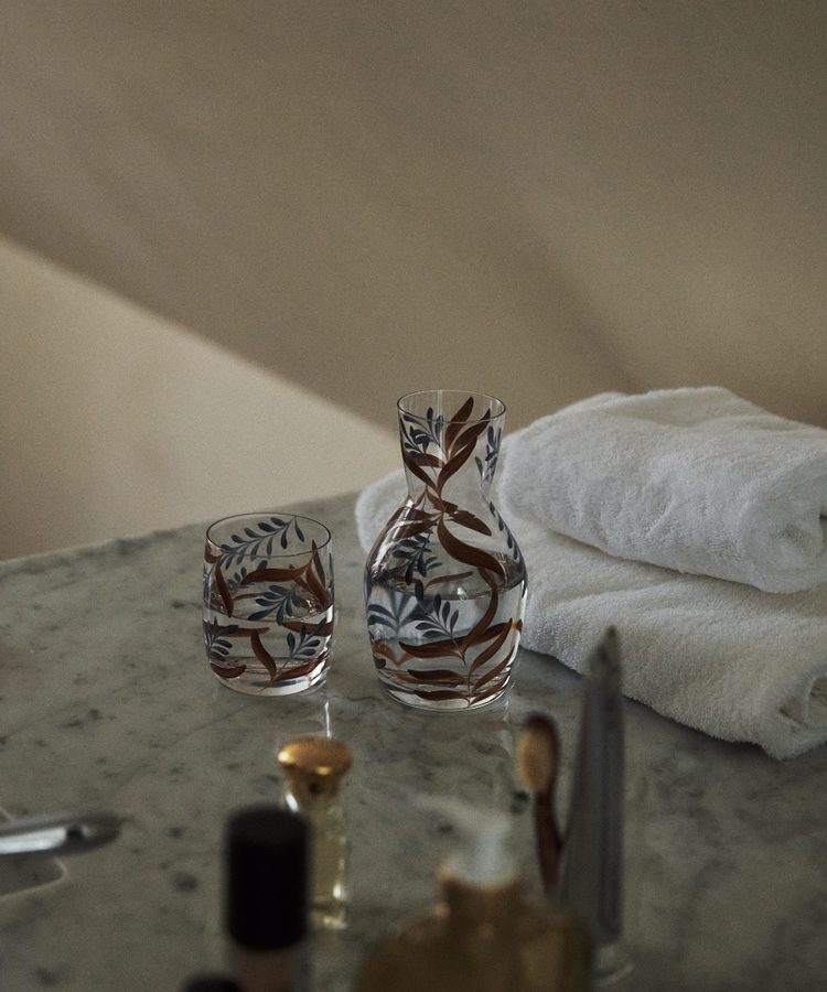 The Botanical Bedside Carafe and Glass Set, featuring a hand-painted botanical design, rests on a marble surface alongside two folded white towels and assorted toiletries.