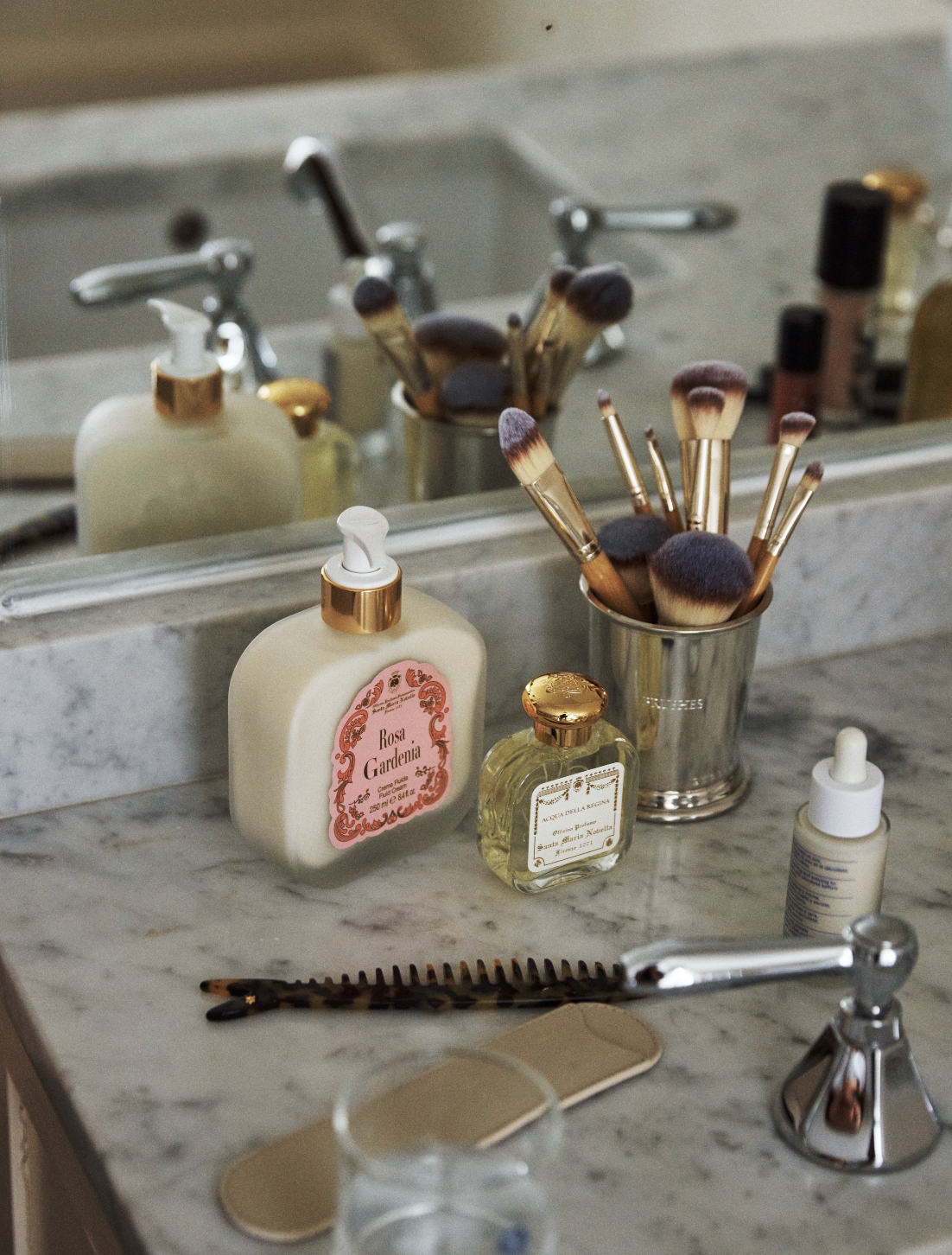 A bathroom countertop features the Jane Brushes Cup filled with makeup brushes, alongside lotion bottles, a comb, a hairbrush, and a faucet in front of a mirror.