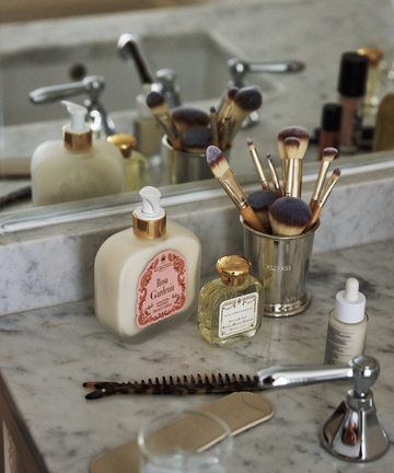 A bathroom countertop features the Jane Brushes Cup filled with makeup brushes, alongside lotion bottles, a comb, a hairbrush, and a faucet in front of a mirror.