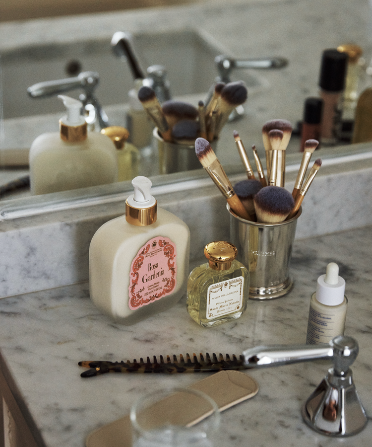 A bathroom countertop features the Jane Brushes Cup filled with makeup brushes, alongside lotion bottles, a comb, a hairbrush, and a faucet in front of a mirror.