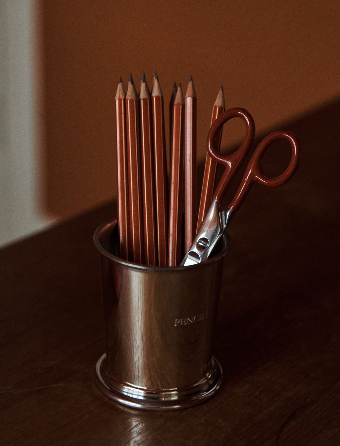 A metal cup holds several sharpened pencils and a pair of Renewal Stainless Steel Scissors with red handles, perfect for everyday use, placed on a wooden surface against a brown background.
