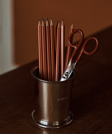A metal cup holds several sharpened pencils and a pair of Renewal Stainless Steel Scissors with red handles, perfect for everyday use, placed on a wooden surface against a brown background.
