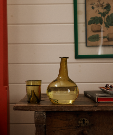 The Elodie Bedside Carafe and Glass Set in yellow, featuring hand-blown glass, sits with water on a wooden table beside a closed notebook; a framed botanical print is displayed on the wall behind them.
