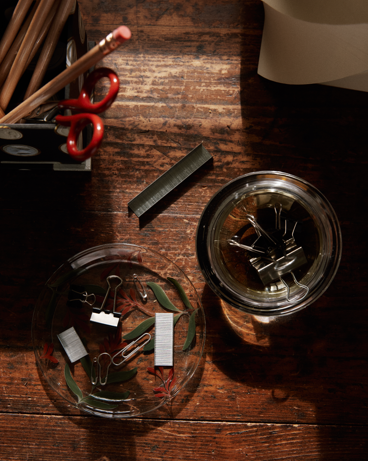 Binder clips and staples in a hand-painted Botanical Little Plate on a wooden desk, with pencils and scissors in a holder nearby.