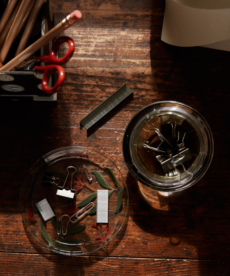 Binder clips and staples in a hand-painted Botanical Little Plate on a wooden desk, with pencils and scissors in a holder nearby.